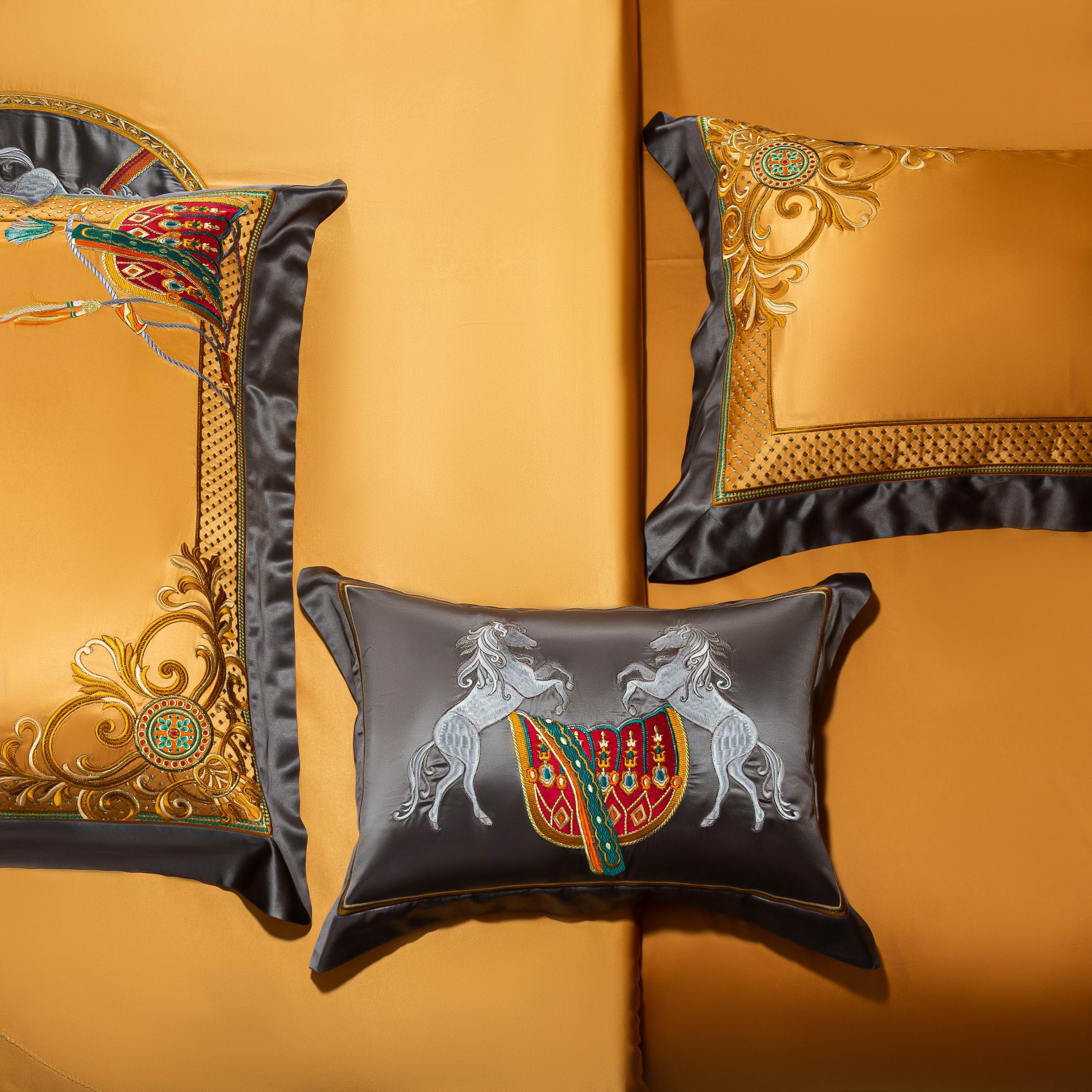 A close-up view of a luxurious golden-yellow bedding set highlighting its intricate design details. The image showcases two decorative pillows: one in black with embroidered white leaping horses and vibrant red and green saddle motifs, and another in gold with ornate baroque-style embroidery, framed with sleek black borders. The duvet corner exhibits matching golden patterns with scrolling accents and a central medallion. The rich contrast of gold and black elements with the detailed embroidery exudes a sen