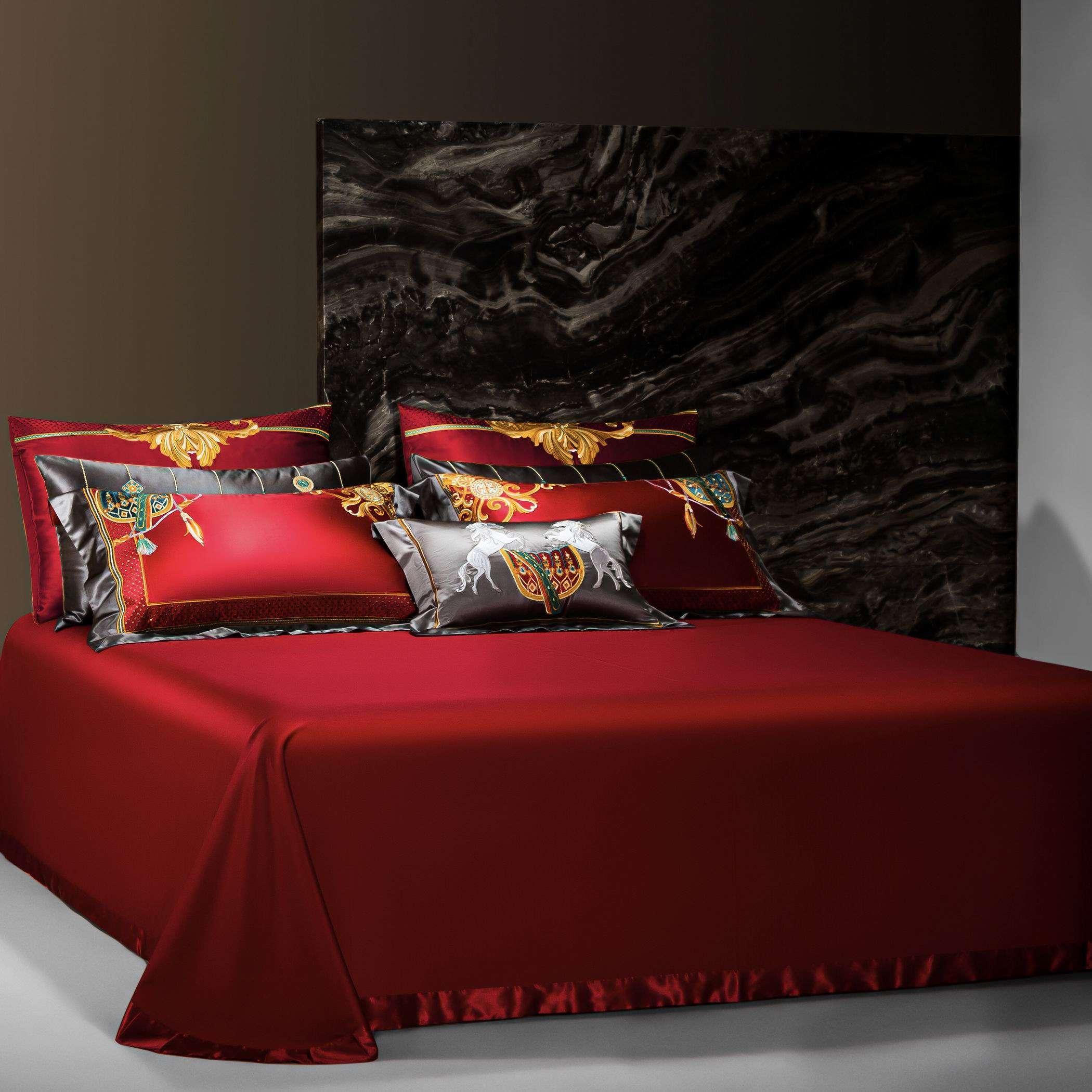 This image features a luxurious red bedding set with a minimalist, smooth base adorned by a set of elaborate, decorative pillows. The backdrop showcases a dark marble accent wall, enhancing the elegance and opulence of the scene. The pillows feature intricate gold and multicolored embroidery, including regal motifs and equestrian elements, adding a striking contrast to the deep red tones. The draping edges of the bedspread emphasize the premium quality of the fabric, exuding a rich and sophisticated aesthet