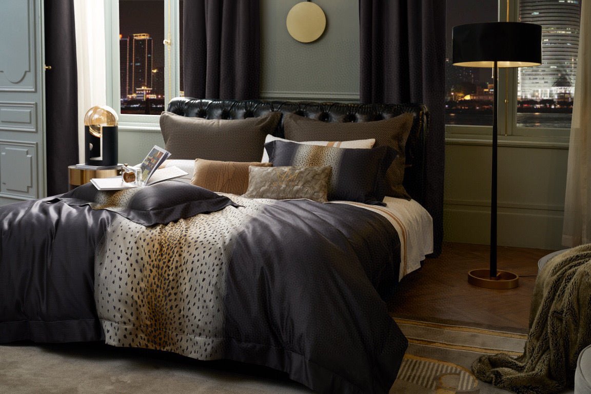Luxurious bedding in deep charcoal tones with a central leopard print design, set in a sophisticated room featuring floor-length curtains, a tufted black leather headboard, and a modern lamp with golden accents.