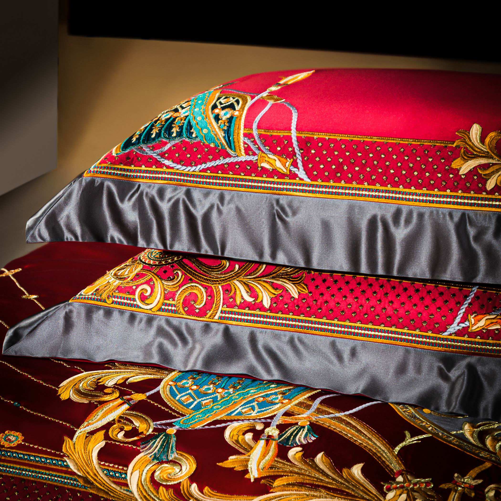 A close-up image of intricately embroidered pillowcases in a deep red satin fabric. The design showcases golden scrollwork, teal accents, and tiny dotted embellishments, creating a luxurious and regal appearance. The pillowcases feature a contrasting gray satin border, adding depth and sophistication to the vibrant red and gold tones. This detailed craftsmanship emphasizes the premium quality and opulent aesthetic of the bedding.