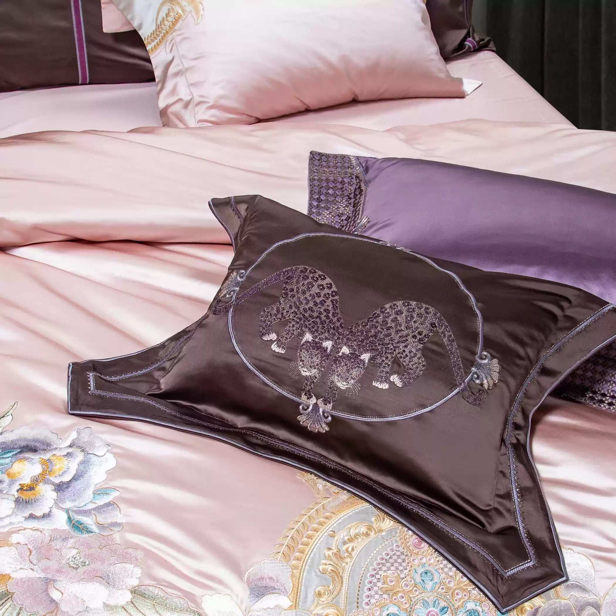 A detailed view of a luxury bedding set featuring a dark brown decorative pillow with an embroidered twin leopard design framed in a circular pattern. The backdrop includes soft pink and lavender silk pillows, accented with floral embroidery and gold embellishments, enhancing the set's opulence and intricate artistry.