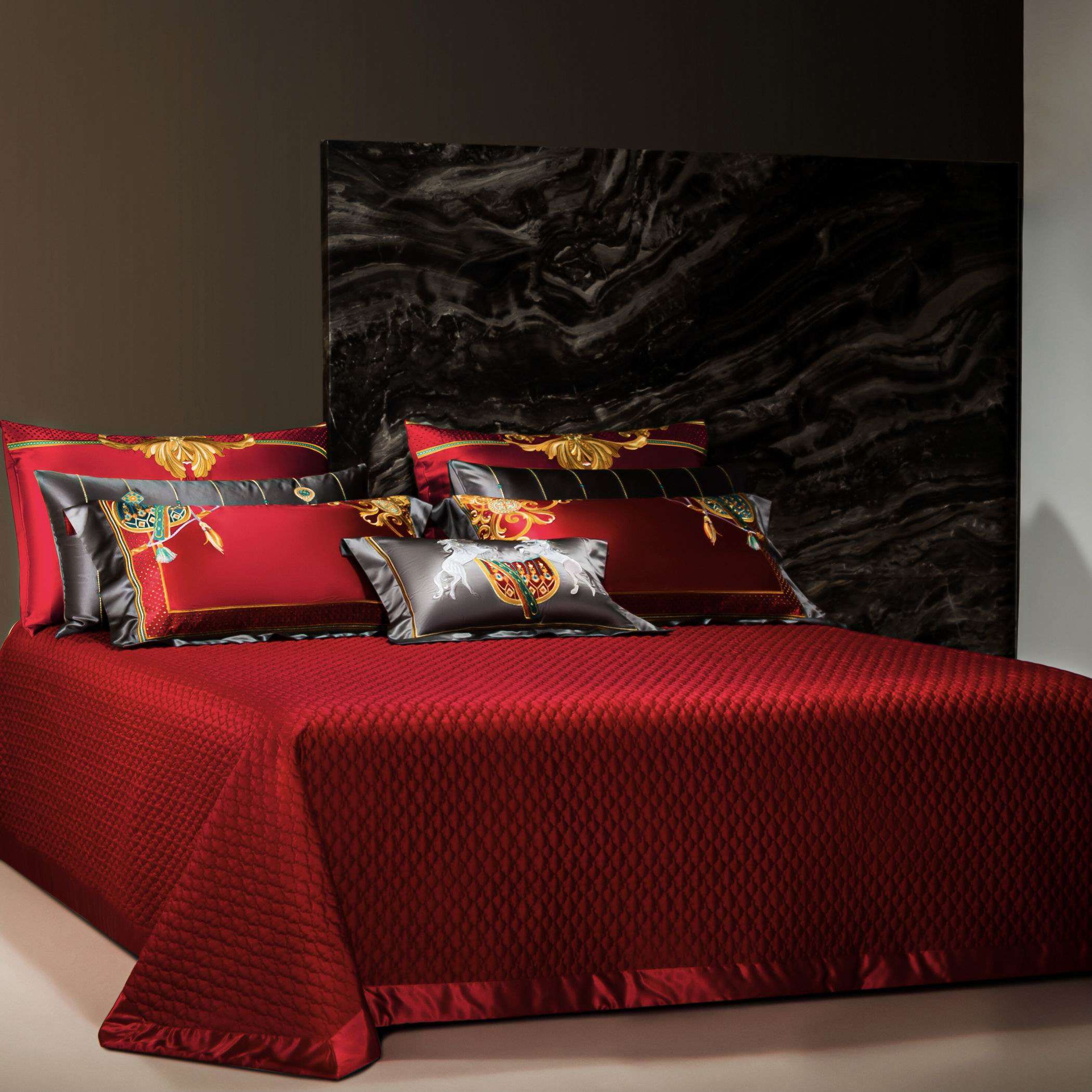 A luxurious red bedding set with a diamond-quilted bedspread draped over the bed, complemented by an array of decorative pillows. The pillows feature ornate gold embroidery, intricate multicolored motifs, and a mix of silky red and black fabrics, adding a regal and elegant touch. The backdrop includes a polished black marble wall, emphasizing the opulence of the bedding design. The smooth, lustrous fabric of the bedspread and pillows reflects a high-quality finish, perfect for a refined and sophisticated be