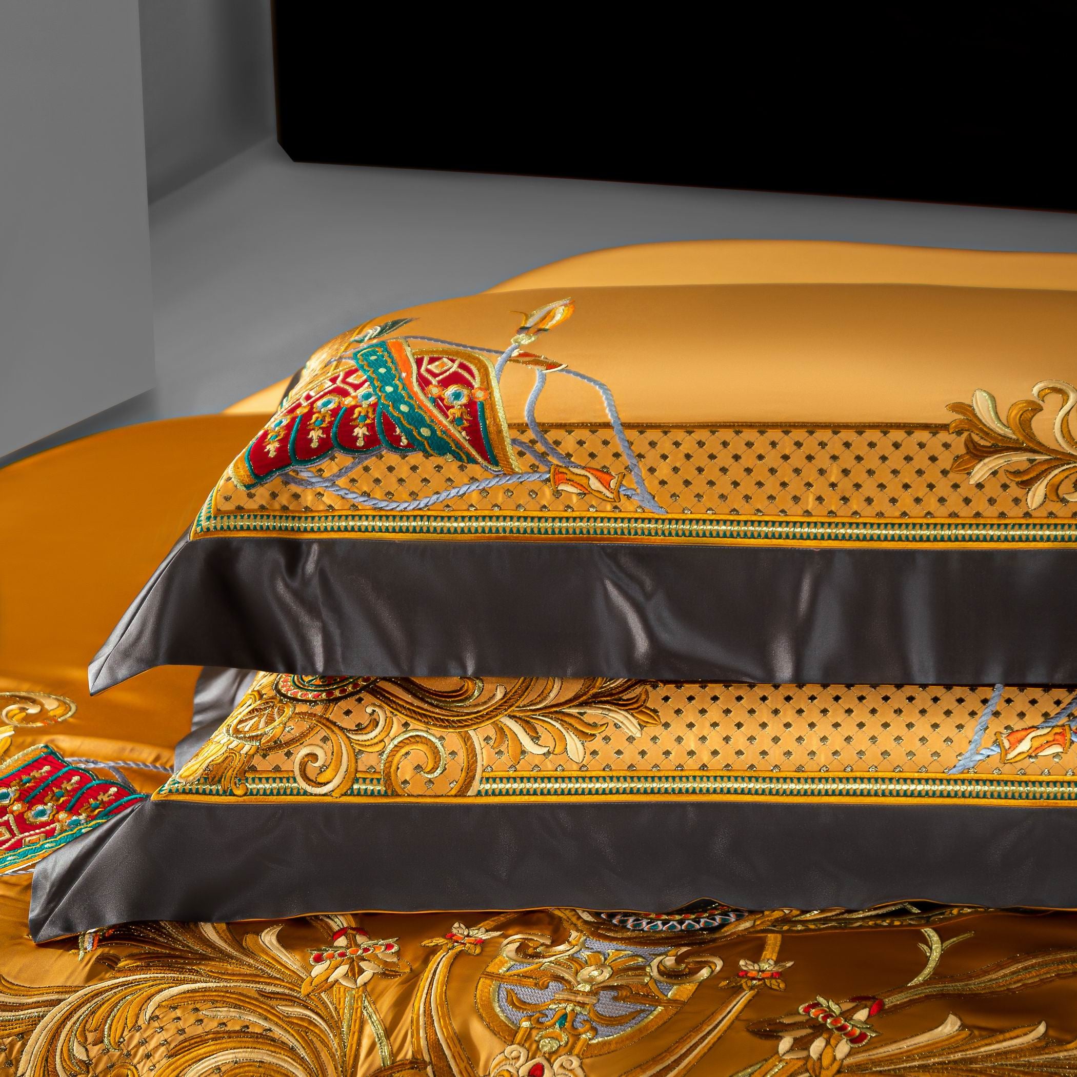 A close-up image of a luxurious golden-yellow bedding set showcasing its elaborate embroidery and detailing. Two stacked pillows are adorned with ornate patterns, including intricate golden swirls, dotted designs, and vibrant red and green saddle-like motifs. The edges are framed with sleek black borders that enhance the elegance and create a striking contrast. Below, the bedding continues the opulent theme with additional baroque-style embellishments, emphasizing the set's lavish and regal aesthetic.