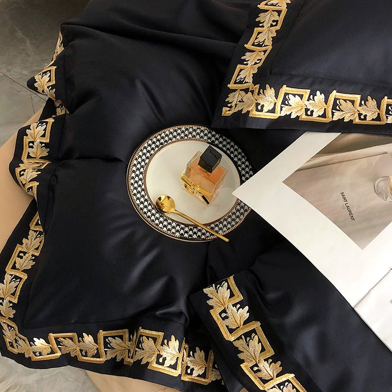 Detailed view of black Egyptian cotton fabric with intricate gold embroidery, complemented by a golden spoon, perfume bottle, and designer plate, showcasing a luxurious and refined aesthetic.