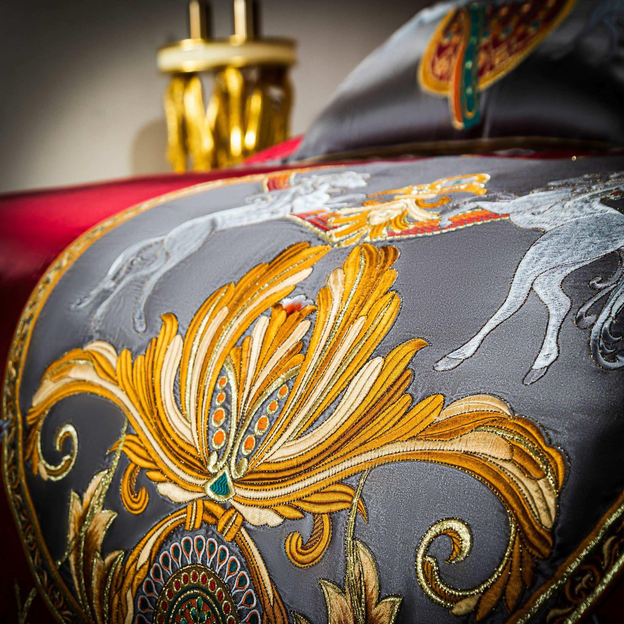 A detailed close-up image of the luxurious embroidery on a bedding set. The design features an ornate golden motif on a gray satin fabric, complemented by intricate stitching in vibrant colors such as teal, red, and orange. Two embroidered white horses are prominently displayed, adding an element of elegance and regality. The rich texture and shimmering fabric emphasize the opulence and meticulous craftsmanship of the bedding. In the background, a golden decorative element subtly enhances the overall grande