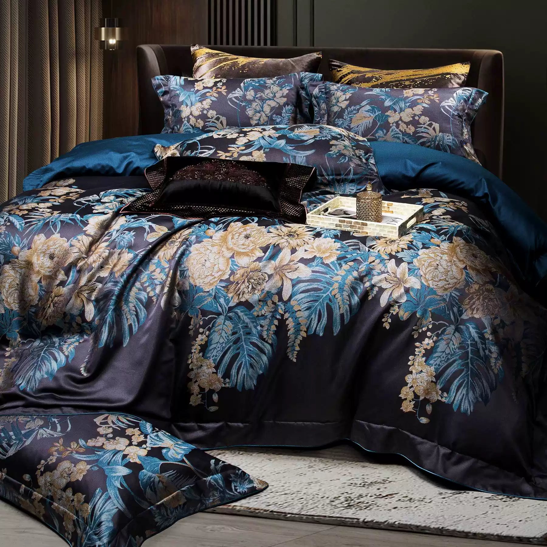 This bedding set continues the luxurious theme with an intricate floral design in vibrant blue and gold tones against a dark backdrop. The composition features a layered arrangement of pillows and a coordinating duvet cover, offering depth and a cohesive aesthetic. The rich teal bedsheet contrasts beautifully with the dark tones, while a black accent pillow with shimmering gold detailing enhances the elegance. The setting is complemented by ambient lighting and a patterned area rug, creating a warm and soph