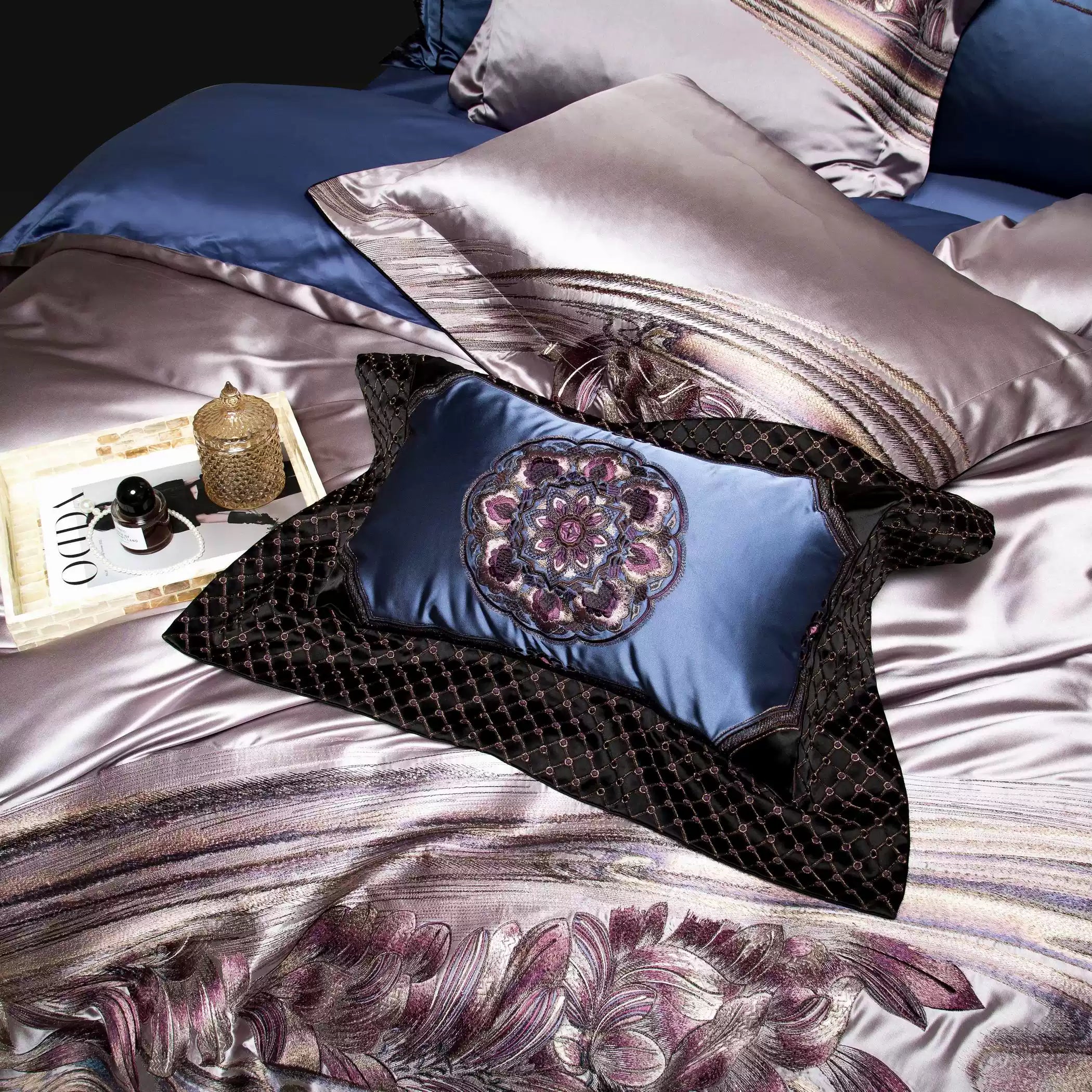 Silky lavender bedding with intricate floral embroidery in shades of purple and pink. The arrangement includes a decorative navy pillow with an embroidered medallion design, accompanied by a matching quilted black overlay. A tray with a perfume bottle and a golden jar is placed on the bedding, adding a luxurious touch.