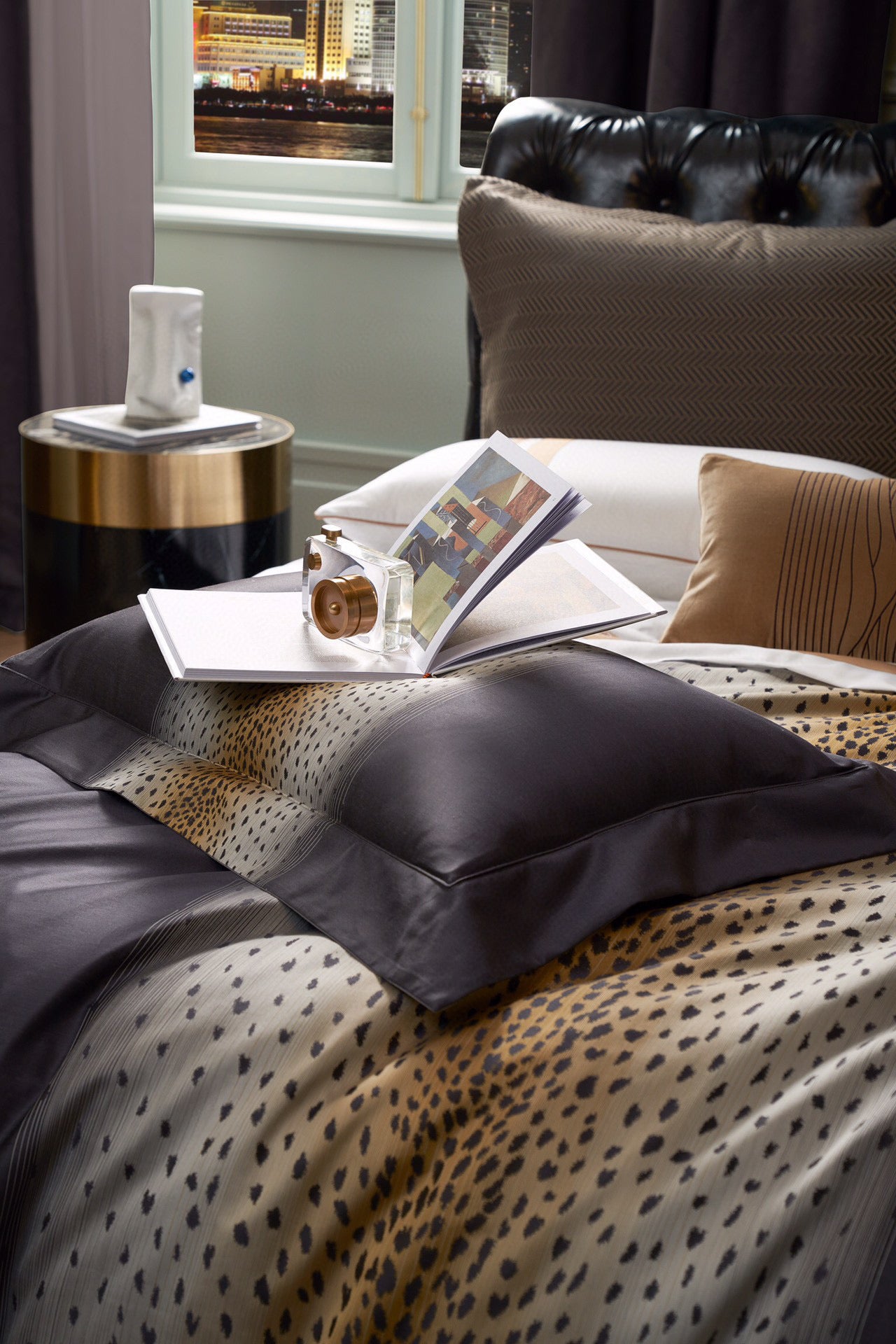 Bedding with a central leopard print gradient and deep charcoal accents, styled with complementary cushions and paired with a modern side table holding a decorative item and book under soft lighting.