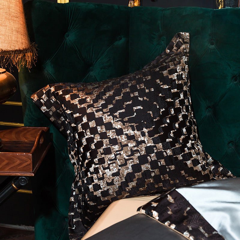 A decorative pillow with a black base, showcasing an abstract geometric pattern in gold and silver tones. The shimmering design creates a textured, luxurious look, perfectly complementing the deep green upholstered headboard. The pillow is made from high-quality Egyptian cotton, adding a smooth, soft finish that elevates its opulence and comfort.