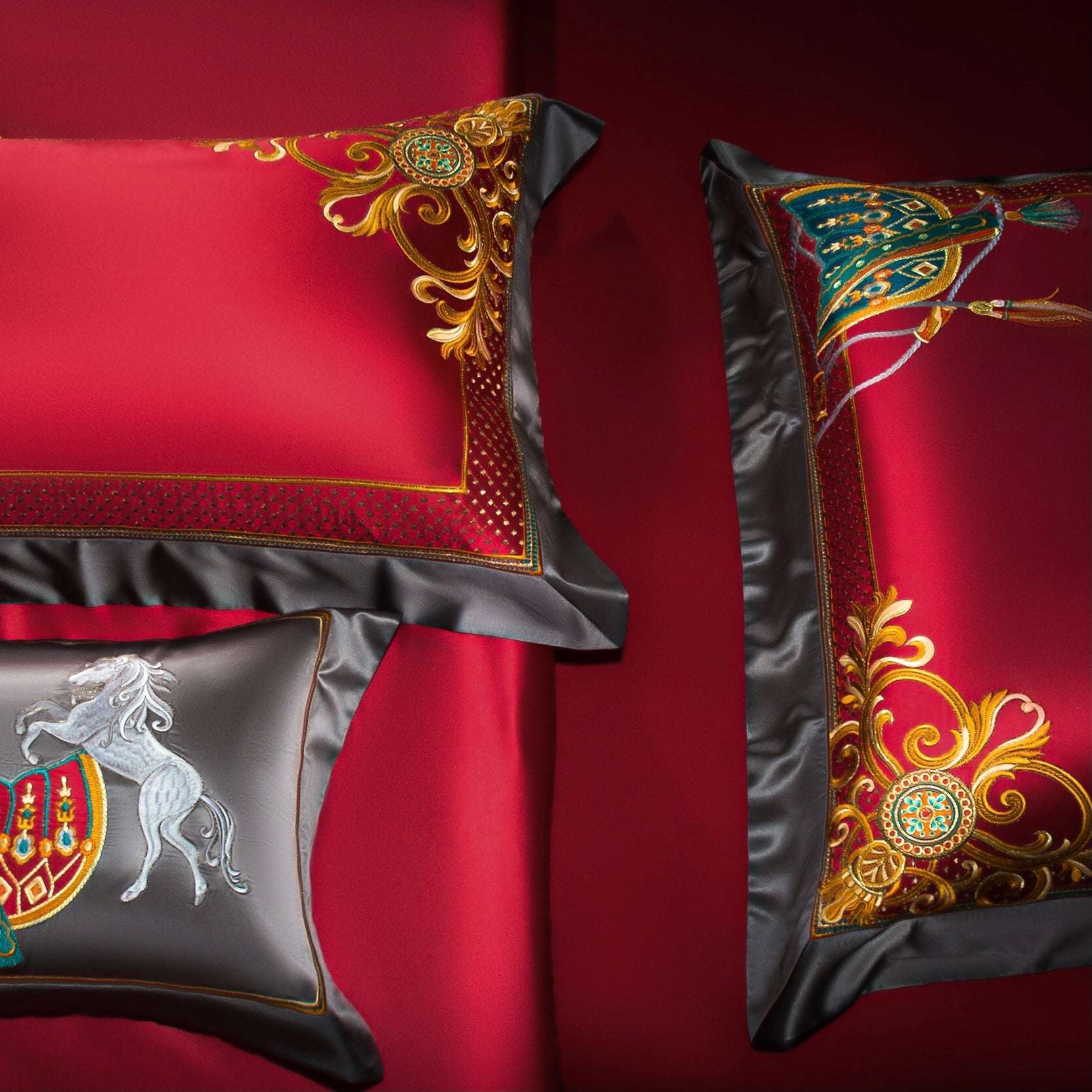 A detailed image of three luxurious pillowcases set against a rich red satin bedding background. The pillowcases feature ornate embroidery in gold, teal, and red, with intricate floral and heraldic motifs. One pillowcase displays a stunning embroidered horse motif on a black satin base, adding a touch of regality. The other pillowcases are adorned with golden swirling patterns and geometric accents, bordered with sleek black satin trims. The vivid contrast between the red and black tones enhances the opulen