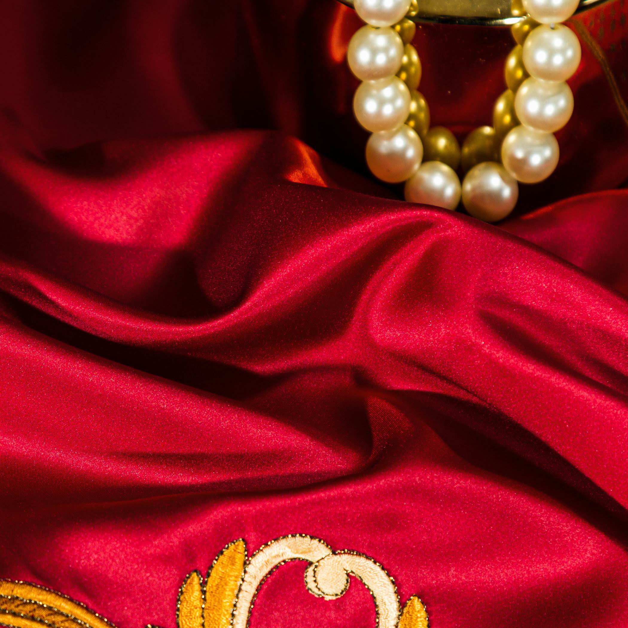 A close-up view of luxurious red satin fabric with a radiant sheen, showcasing its smooth and rich texture. The fabric features a subtle golden embroidery detail, enhancing the opulent aesthetic. In the background, a gold-accented bracelet adorned with lustrous pearls rests elegantly, adding a sophisticated touch to the composition. The lighting highlights the vibrant crimson tones and the reflective quality of the satin, emphasizing its premium craftsmanship.