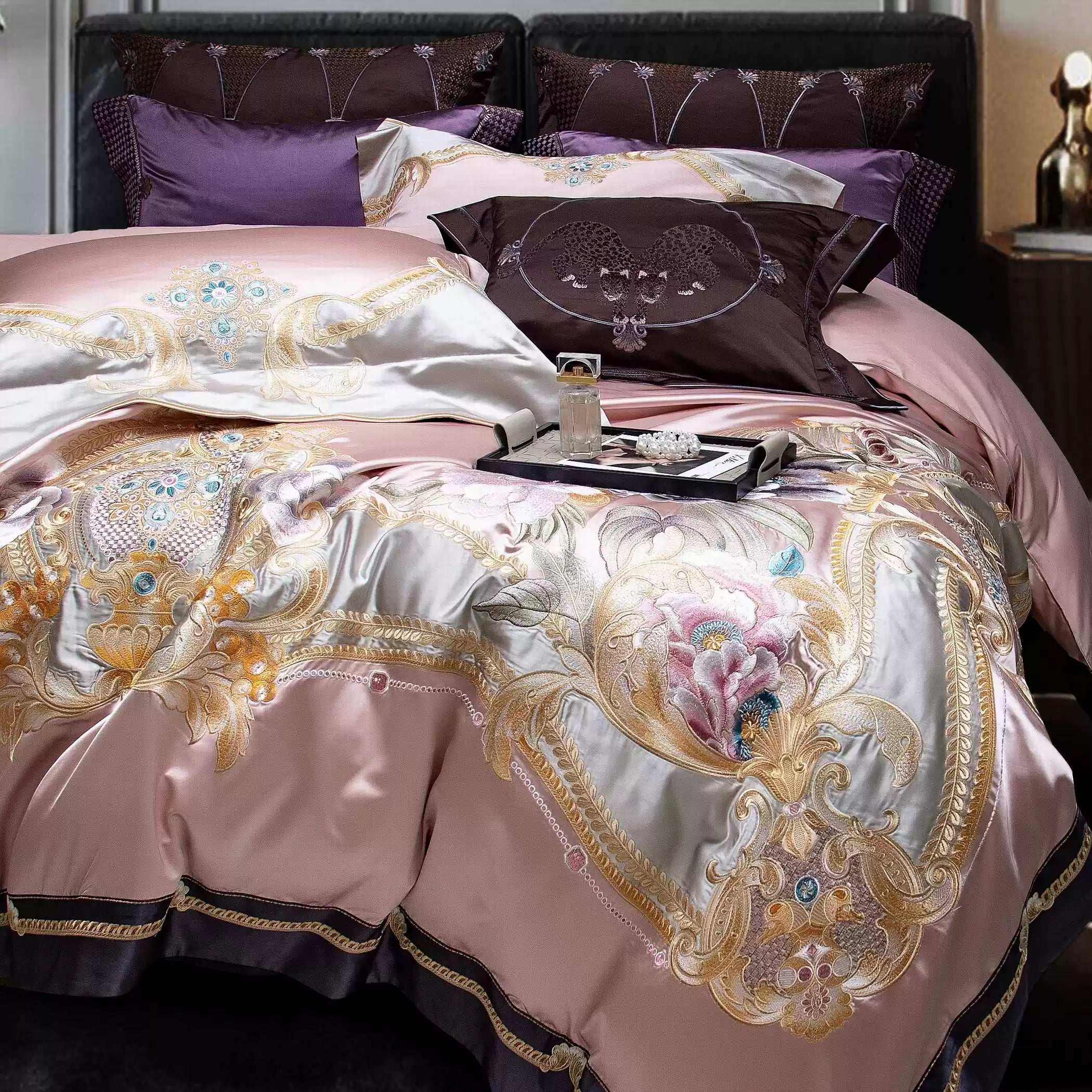 A close-up of an opulent bedding set in blush pink silk, featuring intricate gold embroidery and delicate floral designs. The shimmering accents are complemented by plum and lavender pillows in the background, adding a rich contrast to the luxurious texture and detailing.