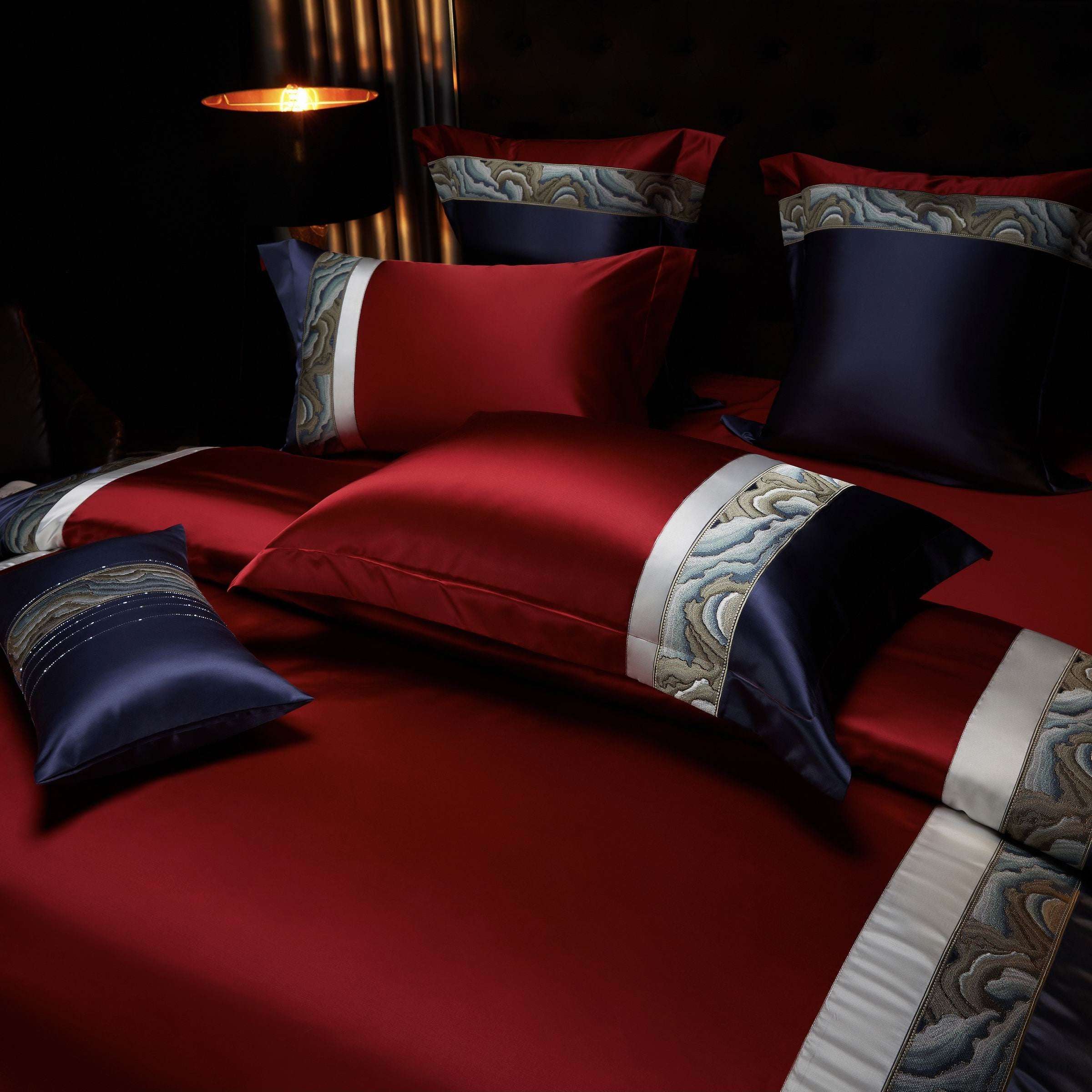 The arrangement highlights the luxurious combination of deep burgundy and navy tones, accented with elegant marble-patterned trims in soft blue and neutral shades. The layered pillowcases and matching bolster pillow create a harmonious aesthetic, complemented by the ambient glow of a nearby lamp. The smooth satin texture and detailed craftsmanship exude sophistication, making it an ideal choice for an upscale bedroom setting.
