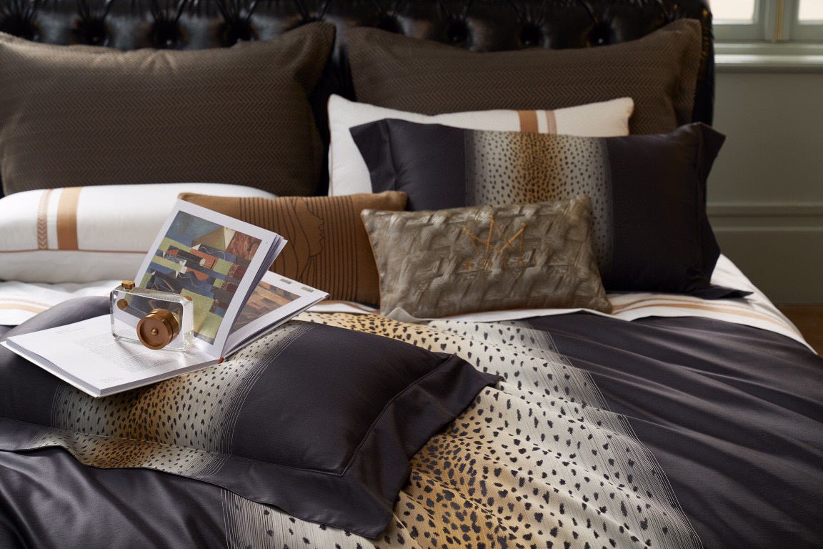 Decorative bedding arrangement with leopard print detailing and complementary throw pillows in various textures and patterns. A book and a modern glass accessory rest atop the duvet, adding a touch of luxury to the dark-toned bed with a tufted leather headboard.