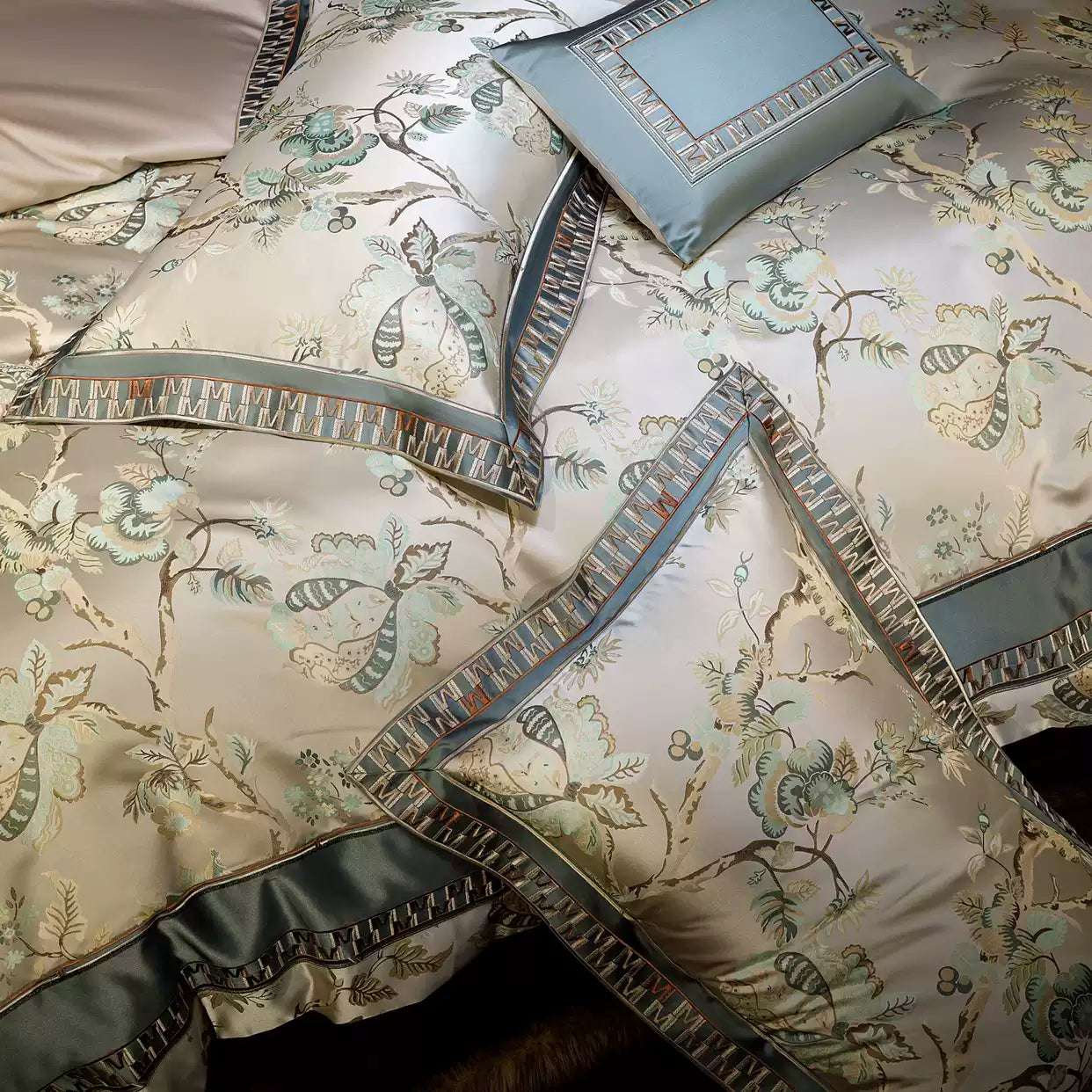 This detailed view highlights the luxurious bedding set adorned with intricate floral and butterfly motifs in soft beige and sage green tones. The silky fabric exudes elegance, enhanced by geometric borders and decorative trims in complementary hues. The pillows display coordinated designs, including a rectangular accent pillow with a bordered frame, adding a sophisticated touch. The fine stitching and lustrous finish of the material emphasize the craftsmanship and premium quality of the set, making it a ce