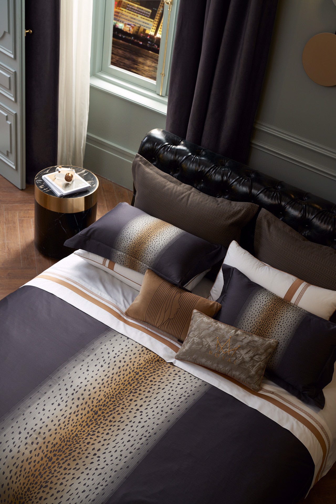 Bedding set in Egyptian cotton featuring a central leopard print gradient design in warm gold and black tones. Paired with coordinating decorative pillows, including embroidered and velvet accents, arranged on a tufted leather headboard. Styled in a sophisticated bedroom with elegant furnishings and a nighttime cityscape visible through large windows.
