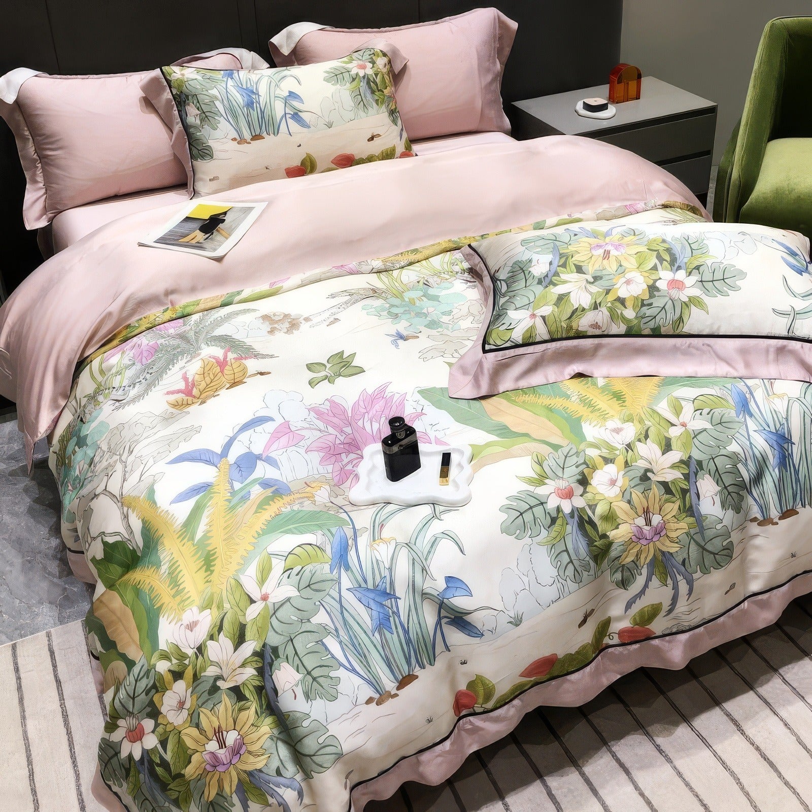 Lyocell eucalyptus fabric bedding set featuring a soft pink duvet cover and a vibrant botanical comforter with detailed floral and greenery patterns in pastel shades. Includes matching pillowcases with floral motifs and pink borders. The scene is styled with a dark grey upholstered headboard, minimalist side tables, a green accent chair, and decorative items like a tray with perfume and accessories, creating an elegant and serene bedroom atmosphere.