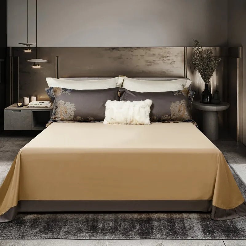 A minimalist bedding arrangement featuring a beige fitted sheet paired with dark gray pillowcases adorned with golden floral patterns. A white faux fur pillow sits at the center, adding texture and contrast. The sleek room design includes a side table with decorative elements and soft, ambient lighting.