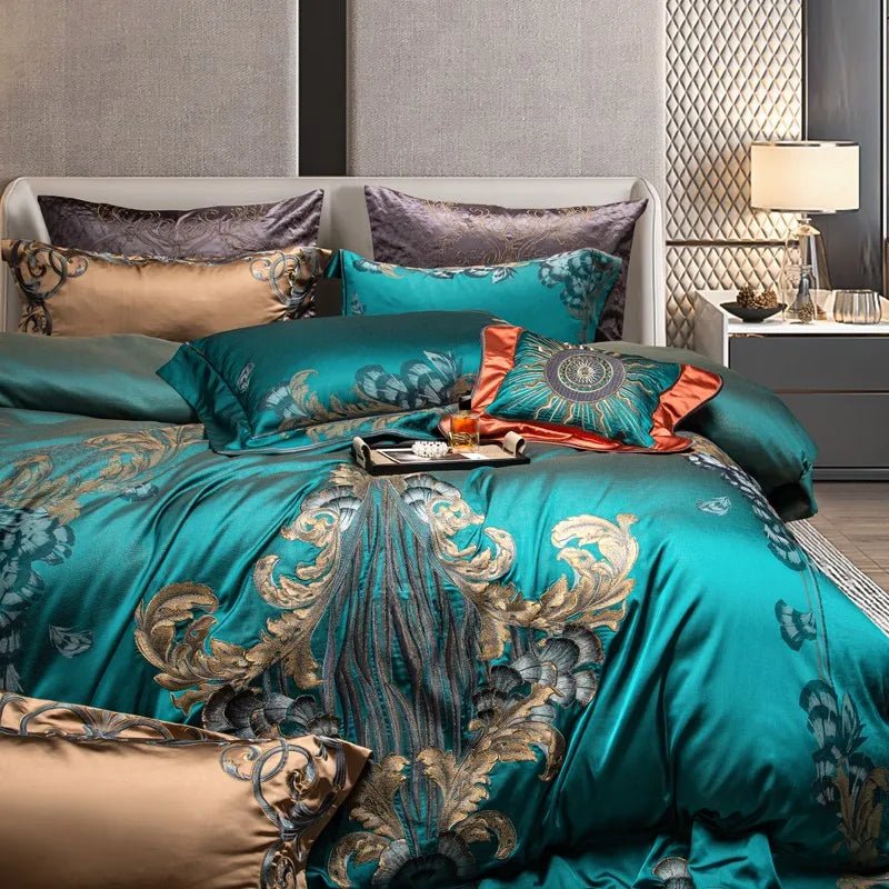 A teal bedding set adorned with intricate baroque-inspired gold and silver embroidery, creating a lavish and sophisticated look. The centerpiece features an eye-catching coral and teal decorative pillow with an ornate sunburst design. The bedding is complemented by coordinating pillows in teal, beige, and gray with embroidered and patterned details. A black tray on the bed holds a gold perfume bottle and a magazine, adding a touch of opulence. Warm bedside lighting and quilted walls in the background enhanc