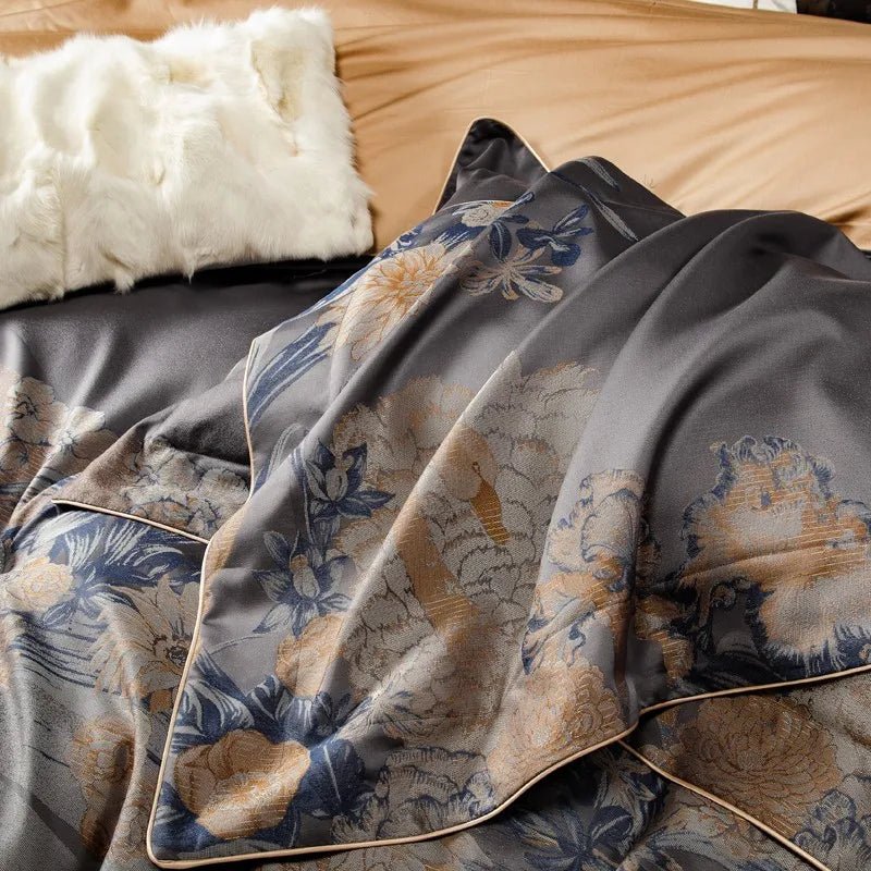 Dark gray jacquard bedding with soft golden and blue floral patterns, accented by a draped blanket and beige piping details. A white faux fur pillow adds a cozy texture to the arrangement against a beige backdrop.