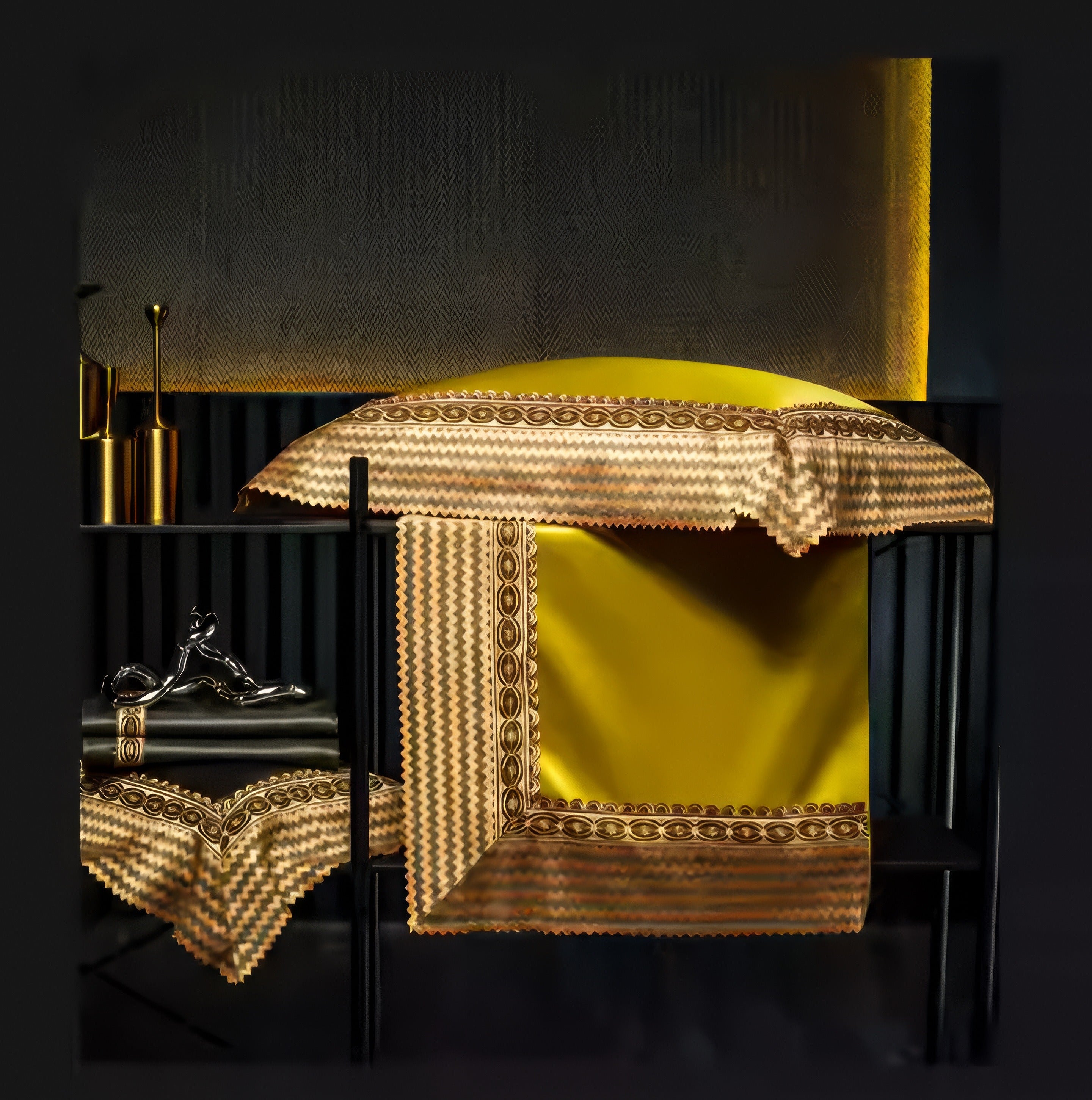 Golden-yellow satin bedding with a decorative zigzag and circular embroidered border draped elegantly over a black bench. A matching folded piece of fabric with the same detailed trim rests on a nearby shelf alongside metallic gold accents and a silver figurine.