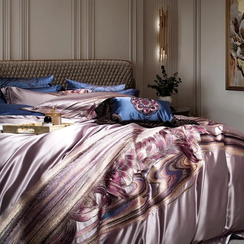 Lavender silk bedding with intricate floral embroidery in shades of purple and pink, complemented by a navy decorative pillow with an embroidered medallion design and a quilted black overlay. A quilted headboard and neutral-toned walls add to the sophisticated bedroom decor. A golden jar and perfume bottle rest on a tray, enhancing the luxurious ambiance.
