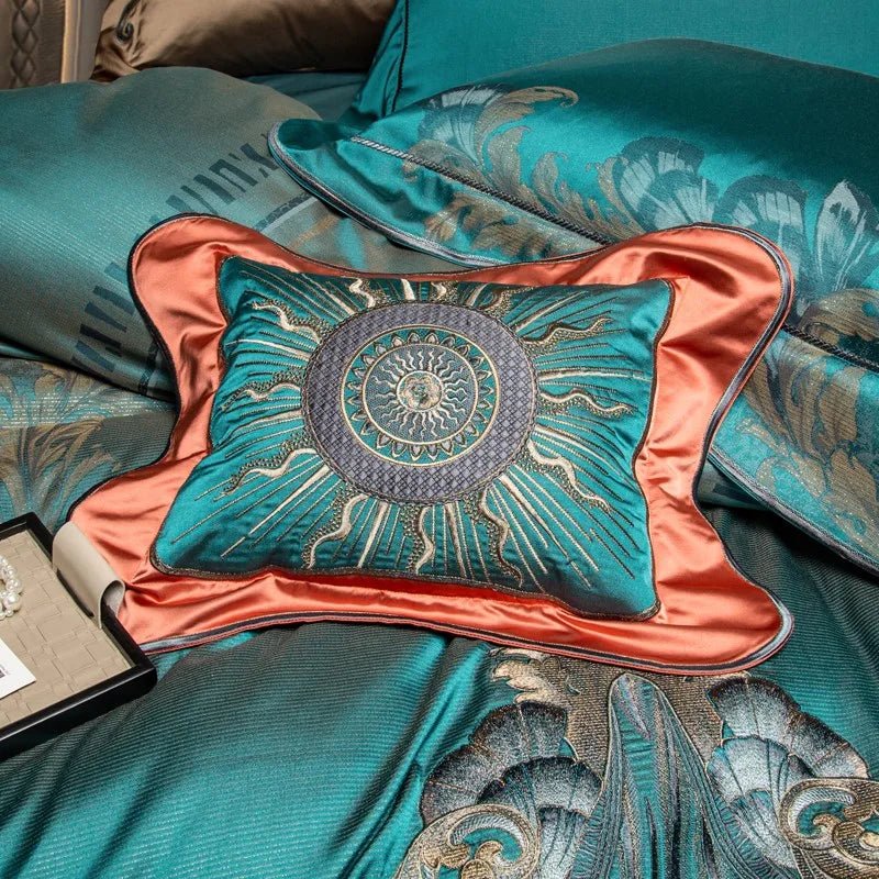 A close-up of a teal pillow with an embroidered sunburst design featuring intricate gold and silver detailing. The pillow is bordered by a contrasting coral satin trim, adding a vibrant accent to the overall luxurious look. Surrounding the pillow are elements of a teal bedding set with elegant baroque-inspired embroidery, blending seamlessly with the design of the decorative pillow.