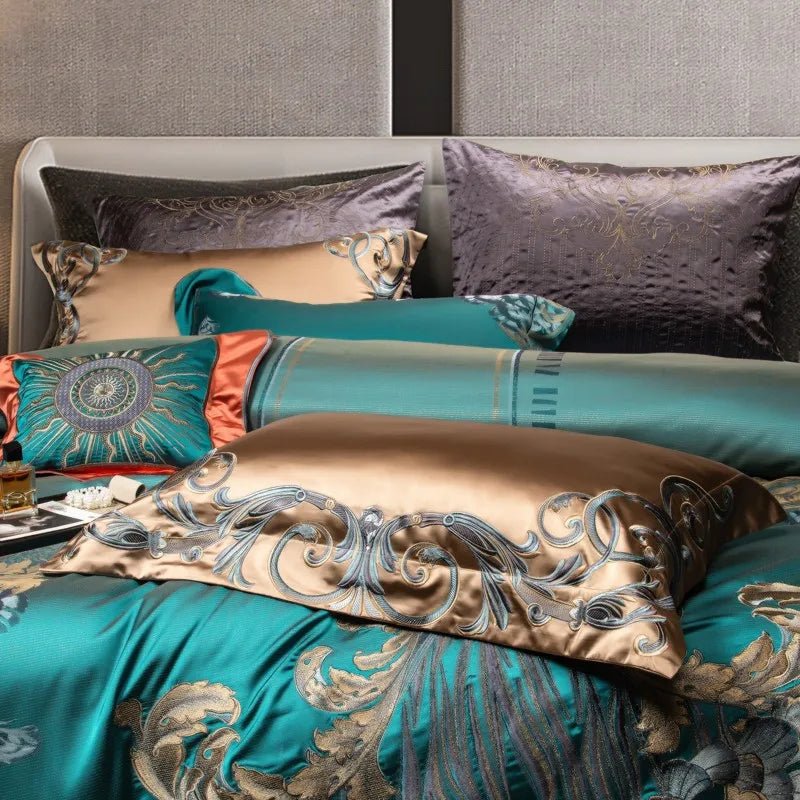A luxurious bedding arrangement showcasing teal and champagne-colored pillowcases with ornate embroidery. The foreground features a champagne pillowcase adorned with intricate swirling patterns in gold and silver thread, complementing the teal duvet with matching baroque-inspired designs. A decorative coral and teal pillow with a sunburst motif adds a bold accent, while textured grey pillows in the background provide depth and contrast. The overall composition exudes elegance and sophistication.