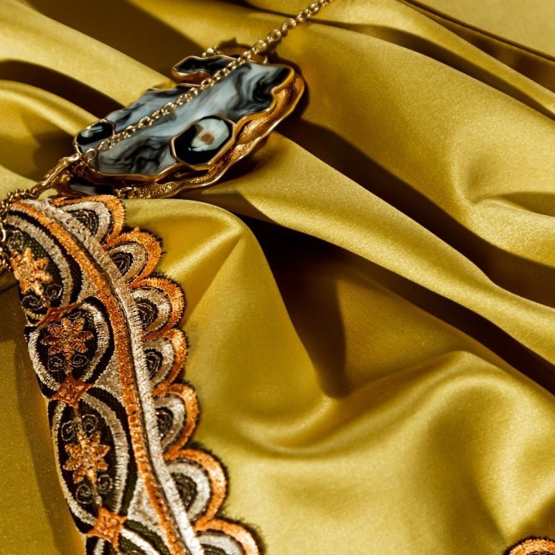 Close-up of a golden-yellow satin fabric with intricate embroidered trim in black, gold, and orange. A decorative gold and black necklace with a marbled pendant lies elegantly on the smooth, lustrous surface of the fabric.