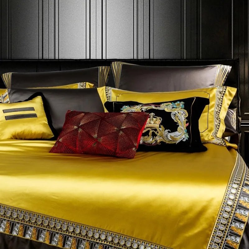 A close-up view of an opulent bedding set featuring a luxurious yellow satin duvet bordered with intricate gold and silver embroidery. The pillows showcase a mix of rich tones: a decorative black velvet pillow with elaborate multicolored embroidery, a red pillow with a textured geometric pattern, and complementary yellow and brown pillows with gold accents. The sophisticated design, combined with the bold color palette, creates an elegant and regal bedroom ambiance.