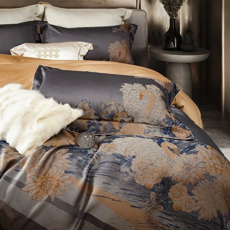 A luxurious bedding set with dark gray jacquard fabric featuring intricate swan and floral patterns in soft golden and blue tones. The arrangement includes accent pillows, a white faux fur pillow, and warm beige undertones, complemented by a modern bedside table and vase in the background.