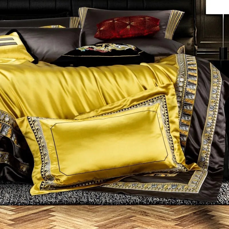 A detailed view of a premium bedding set, featuring a striking yellow pillowcase with a glossy satin finish, bordered by intricate gold and silver embroidery. The pillow rests against a complementary brown duvet cover, accented with matching embroidered trims for a luxurious touch. Behind it, a decorative black pillow with a colorful embroidered design and a deep red textured pillow enhance the rich and regal aesthetic.