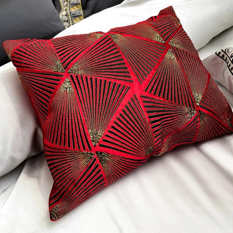 A vibrant decorative pillow in bold red with an intricate geometric pattern of gold and black radiating lines, forming a stunning, fan-like design. The textured fabric enhances the luxurious appeal, making it a striking accent piece against the neutral tones of the bedding.