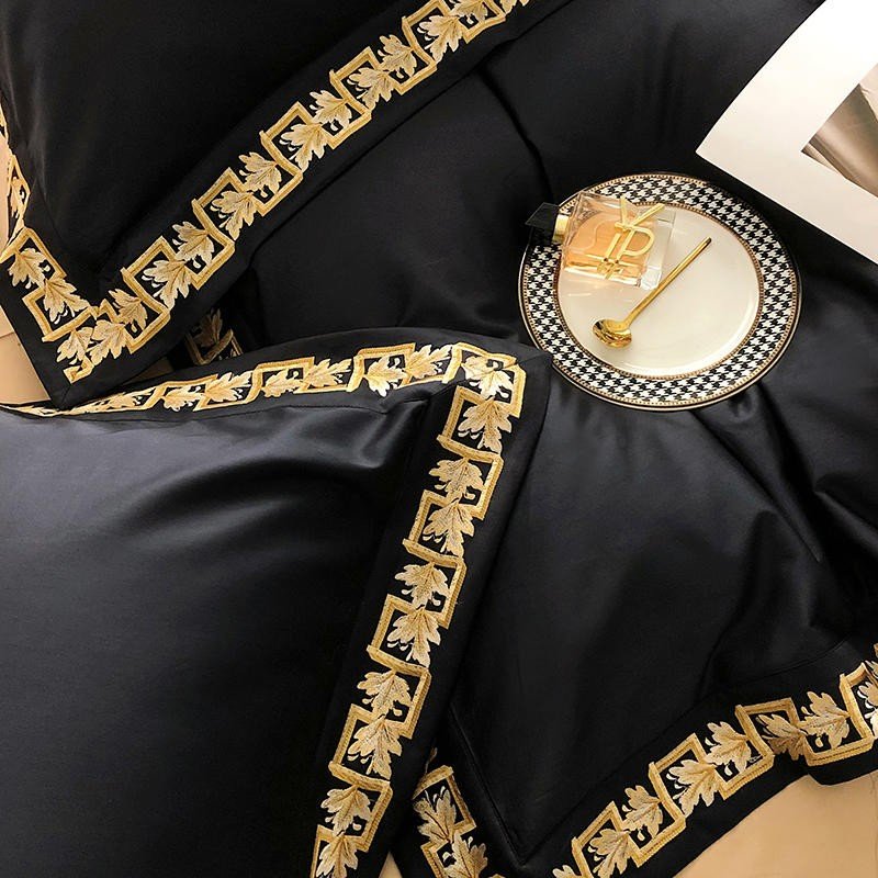 Black Egyptian cotton bedding with gold embroidery detail, accompanied by a designer plate, golden spoon, and perfume bottle for a luxurious ambiance.