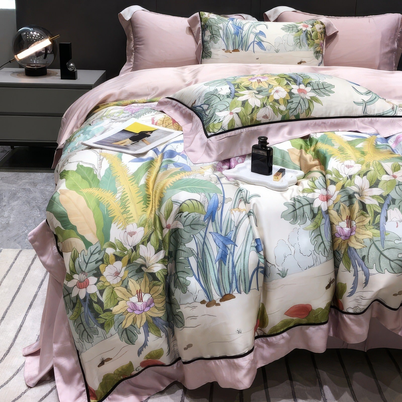 Bedding set made from lyocell eucalyptus fabric, showcasing a soft pink duvet cover and a vibrant botanical-patterned comforter adorned with lush green foliage and colorful flowers. Includes coordinating pillowcases with detailed floral designs and pink borders. The scene features a grey upholstered headboard, minimalist side tables with modern decor, a striped rug, and a tray with a perfume bottle and book, adding a touch of elegance to the setting.