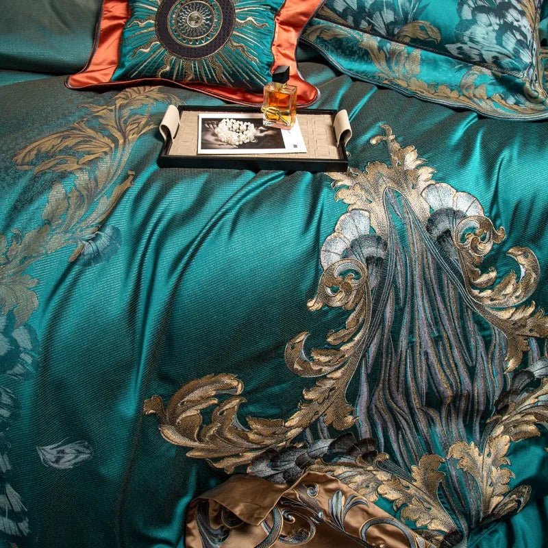 A detailed view of a teal bedding set showcasing intricate gold and silver embroidery in a baroque-inspired design. The flowing patterns and floral accents add depth and elegance to the fabric. A black tray sits on the bedding, holding a gold perfume bottle, a strand of pearls, and a monochrome photograph, creating an opulent display. A decorative pillow with a teal and coral sunburst design is visible in the background, enhancing the luxurious aesthetic.