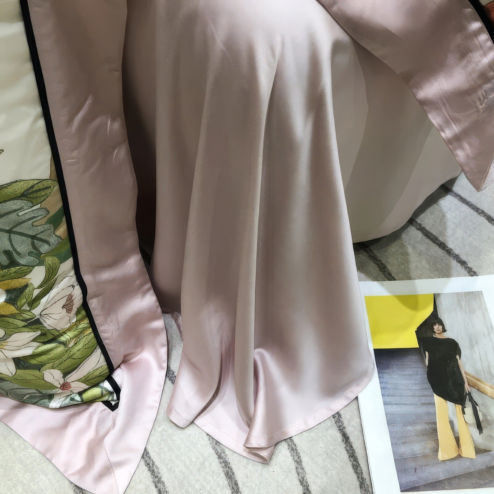 A close-up view of the corner of a luxury Lyocell eucalyptus bedding set, showcasing a blush pink sheet with a soft, silky texture draped elegantly. The edge of a floral-patterned duvet with blush satin trim and black piping is visible, adding a refined touch. A magazine featuring a fashion image is placed on a striped rug nearby, enhancing the scene's modern and artistic ambiance.
