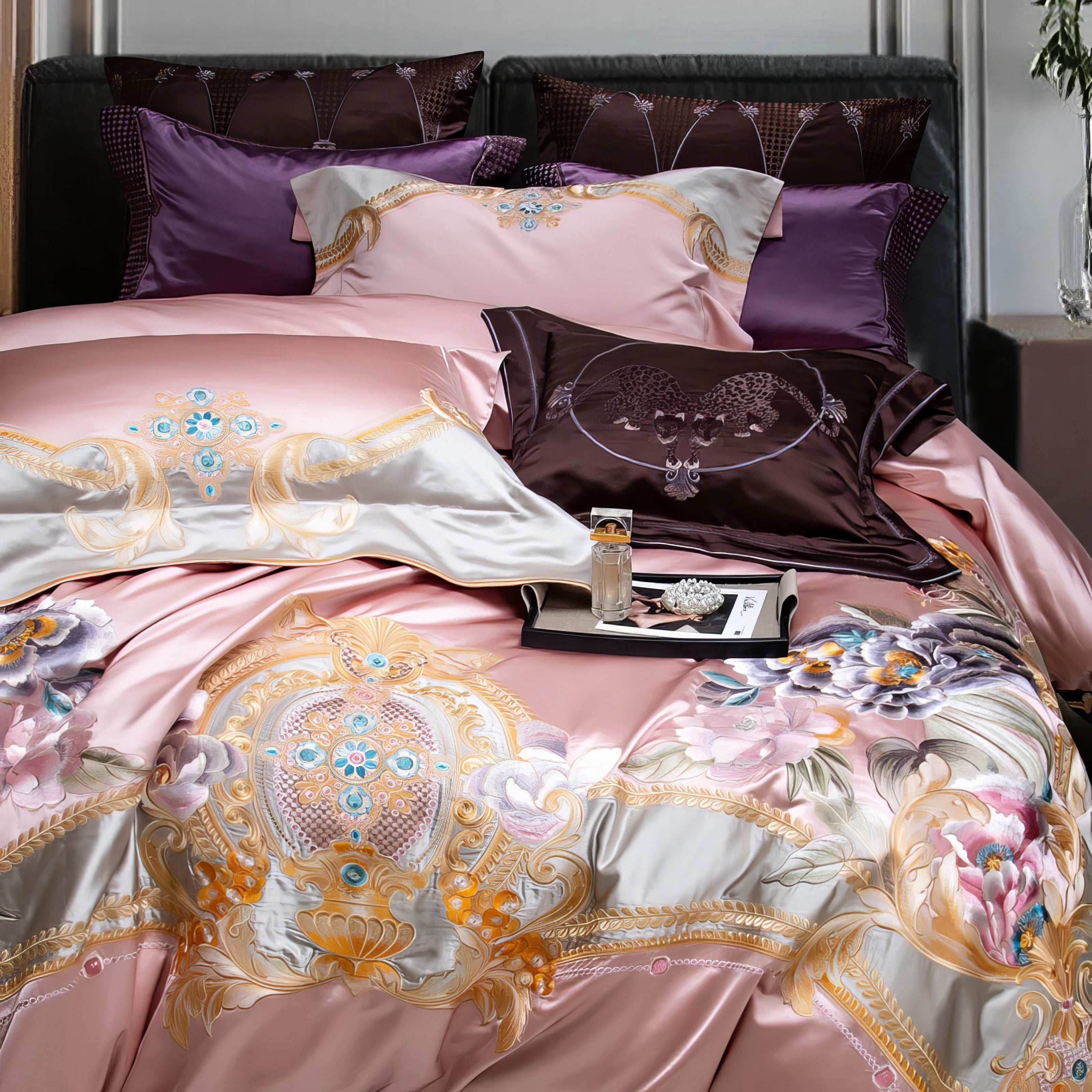 A luxury bedding set in soft blush pink, showcasing intricate gold and pastel floral embroidery. The design is accented with complementary plum and lavender pillows, featuring unique embroidered patterns, adding elegance and sophistication to the setup. A tray with decorative items is placed atop the bed, enhancing its opulent aesthetic.