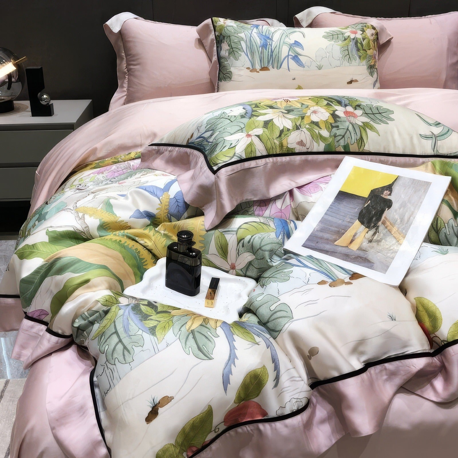 A close-up view of a luxurious bedding set made from eco-friendly Lyocell eucalyptus fabric, showcasing a nature-inspired floral and botanical print with vibrant green, pink, and yellow tones. The bedding includes a duvet cover, pillowcases, and decorative pillows edged with a satin trim in blush pink. A tray holding a black perfume bottle, a lipstick, and a magazine accentuates the sophisticated design, resting atop the quilted comforter.