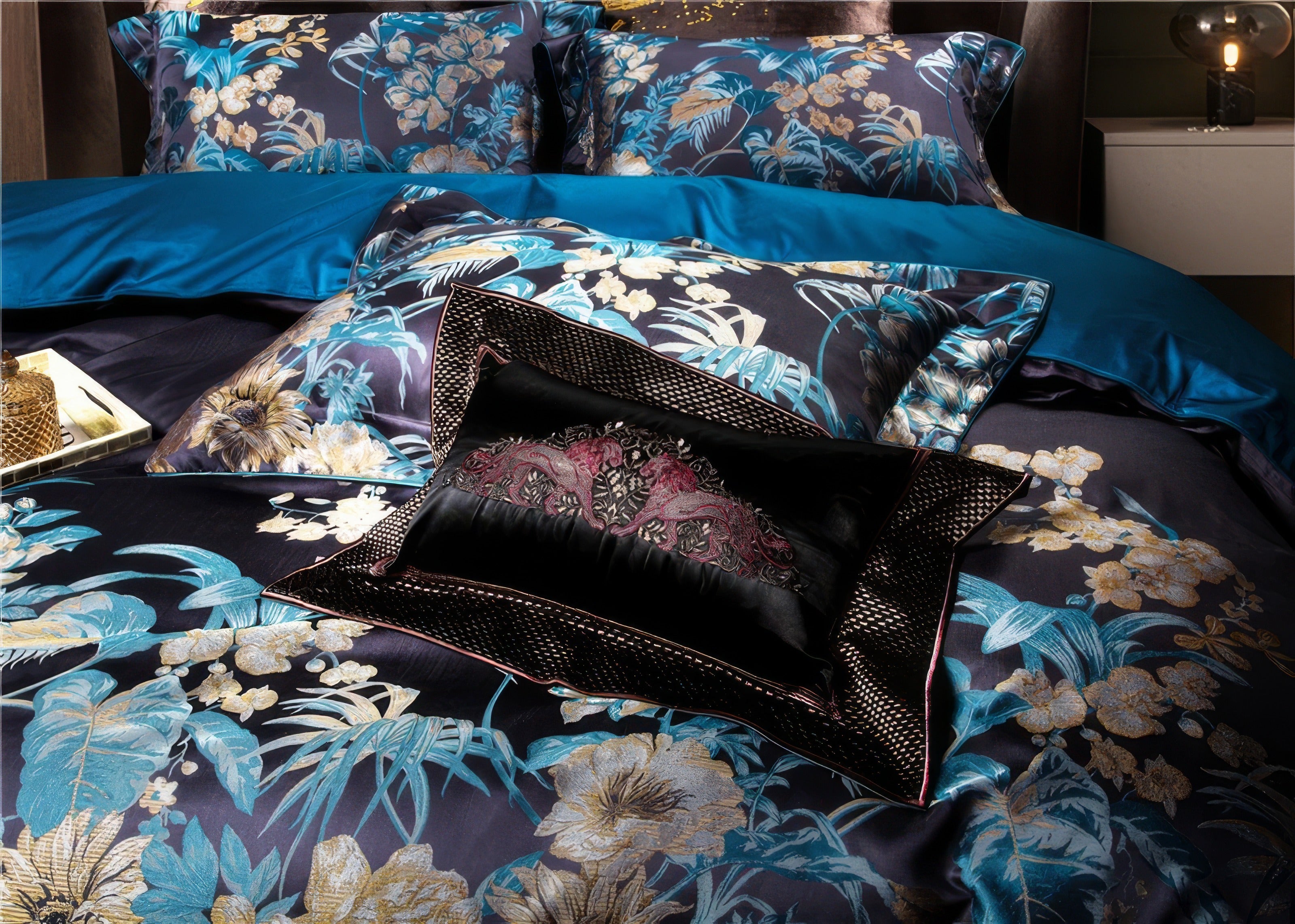 This close-up highlights the intricate craftsmanship of the bedding set. The centerpiece, a black velvet throw pillow with rich, embroidered details in burgundy and gold, exudes elegance and sophistication. Surrounding it, the satin pillowcases and duvet showcase a bold floral motif in shades of teal, gold, and cream, set against a deep navy background. The teal bedsheet adds a vibrant pop of color, while subtle metallic accents on the embroidery elevate the luxurious aesthetic. This arrangement exemplifies