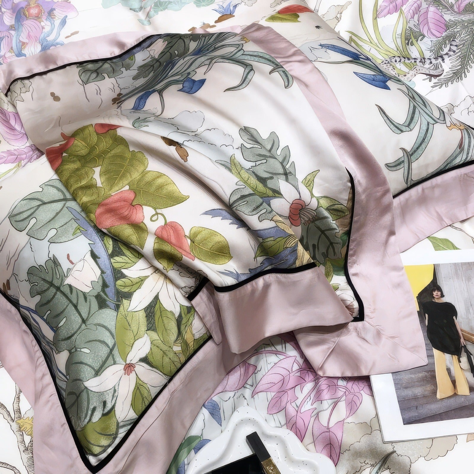 A detailed close-up of a Lyocell eucalyptus fabric pillowcase from a luxury bedding set, featuring a nature-inspired print with leaves, flowers, and soft pastel tones. The pillowcase is bordered with blush pink satin trim and black piping for an elegant finish. The soft and breathable fabric highlights eco-friendly luxury. A magazine and decorative tray with a lipstick rest nearby, adding a stylish accent to the overall composition.