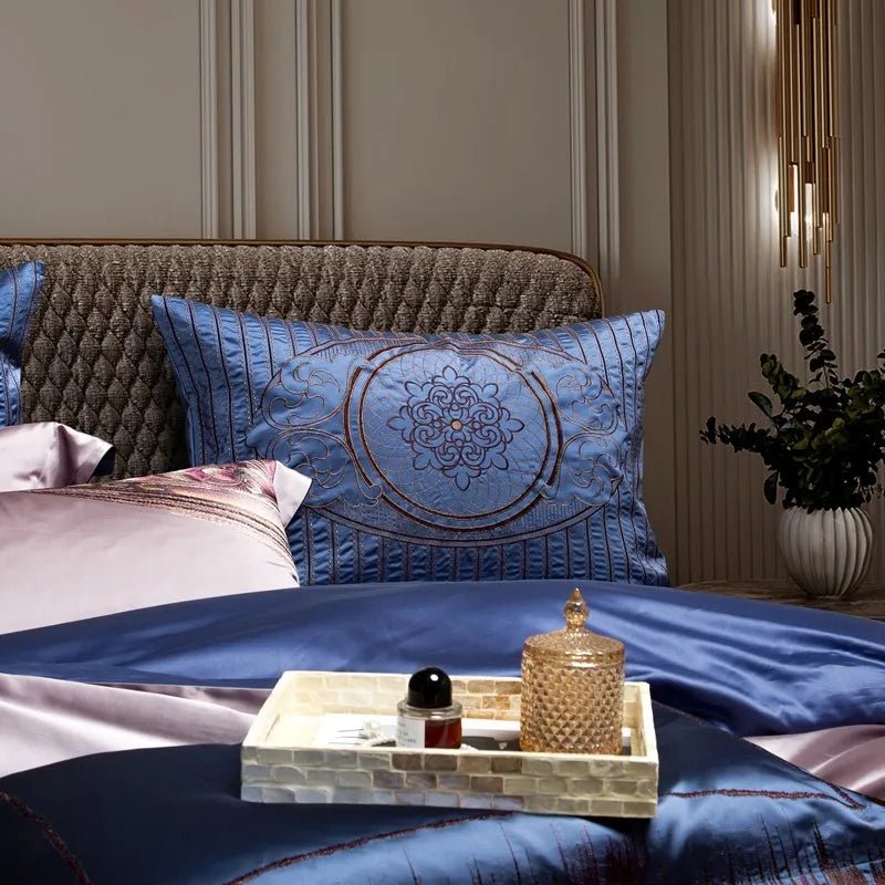 A close-up of a luxurious blue pillowcase with intricate embroidery in the center, paired with silky lavender and blue bedding. A mosaic-style tray on the bed holds a glass jar and a small perfume bottle. The textured headboard and a decorative vase with greenery add a refined touch to the background.