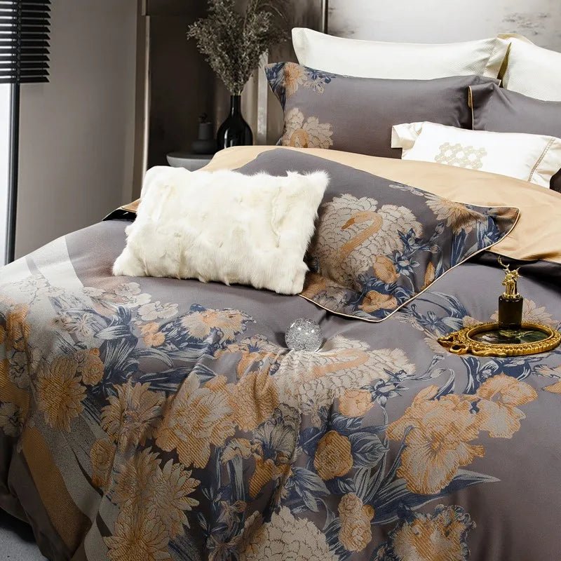 A close-up view of a sophisticated bedding set in dark gray adorned with intricate golden floral patterns and swan motifs. The design is complemented by a cream flat sheet and a plush white faux fur accent pillow, adding texture and elegance. A decorative gold tray holds a perfume bottle and an ornate mirror, alongside a sparkling crystal orb, creating a luxurious and refined ambiance. The background features a modern vase with dried branches and subtle lighting, enhancing the overall contemporary aesthetic