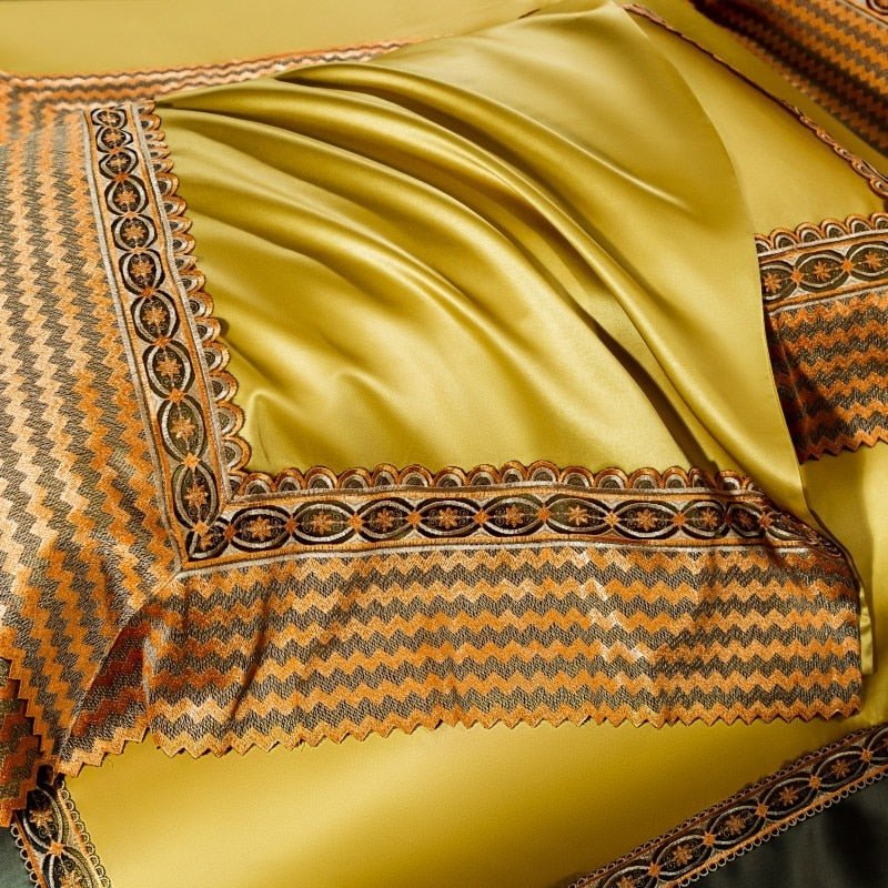 Golden-yellow satin bedding with a smooth, glossy finish and a zigzag embroidered border featuring intricate circular patterns in orange, black, and gold. The fabric drapes softly, showcasing its luxurious sheen and detailed trim.