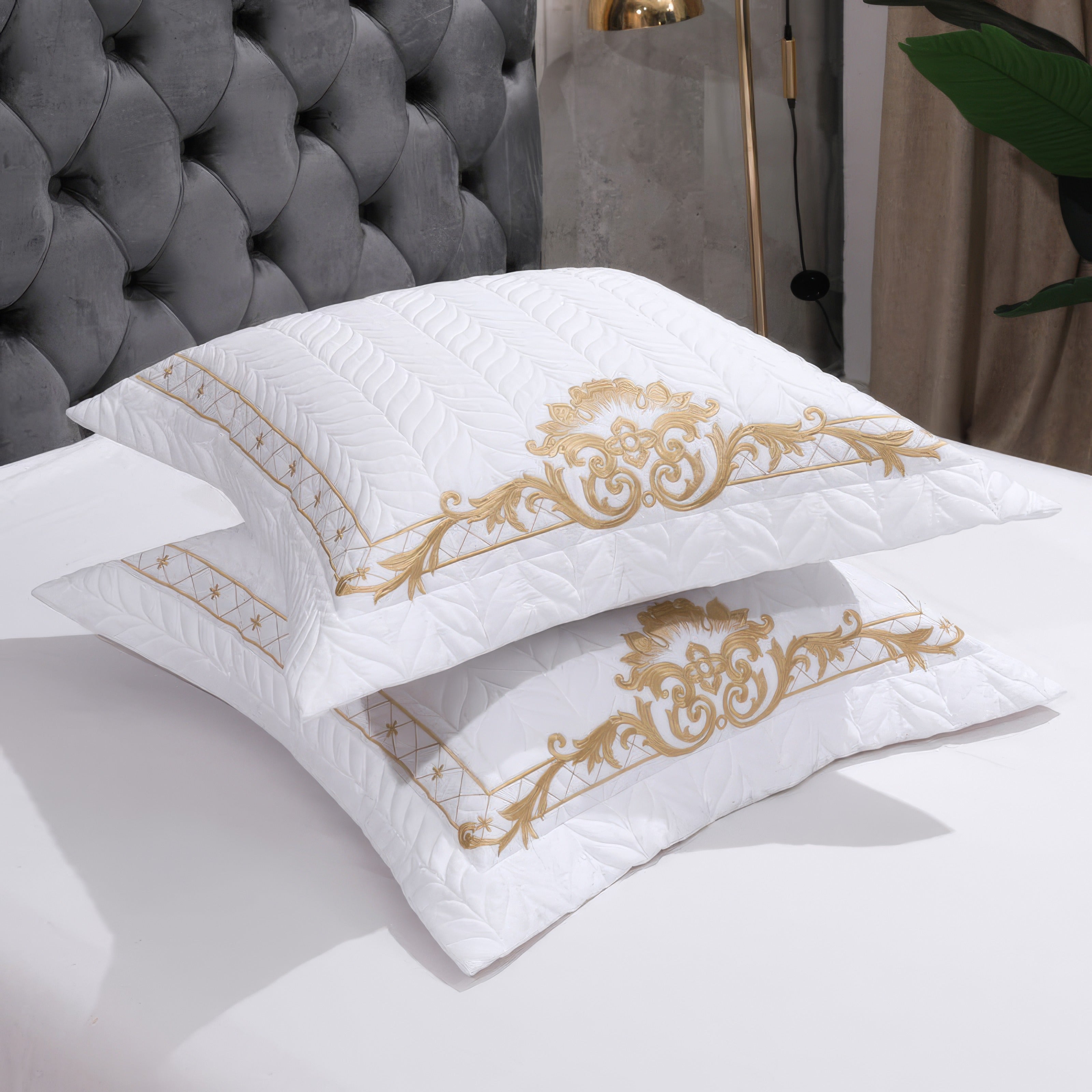 Two white quilted pillows featuring intricate gold embroidery, elegantly designed with baroque-inspired motifs. The tufted gray headboard in the background enhances the luxurious appeal, while the soft white bedding complements the pillows' refined detailing.