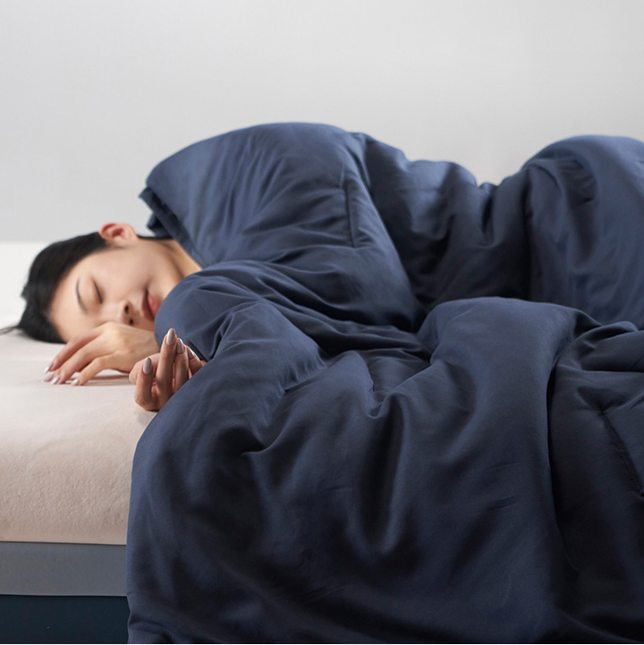 Person sleeping under a blue quilt on a bed with a plain background