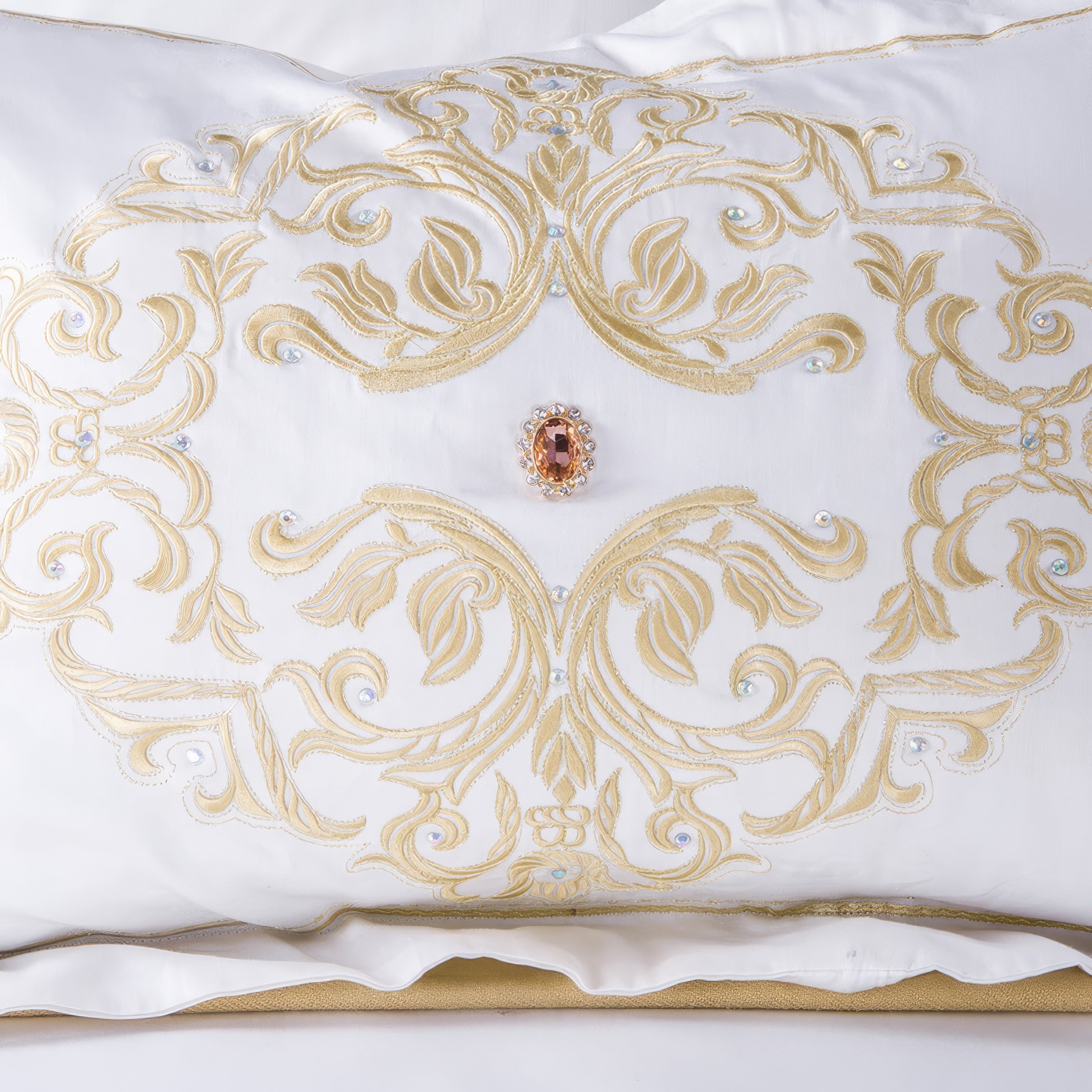 An elegant white pillow with intricate gold embroidery forming a symmetrical baroque-inspired design. The centerpiece features a sparkling jewel accent in a warm amber tone, encased in a delicate frame. The detailing is further enhanced by small crystal embellishments, adding a touch of sophistication and luxury to the overall design.