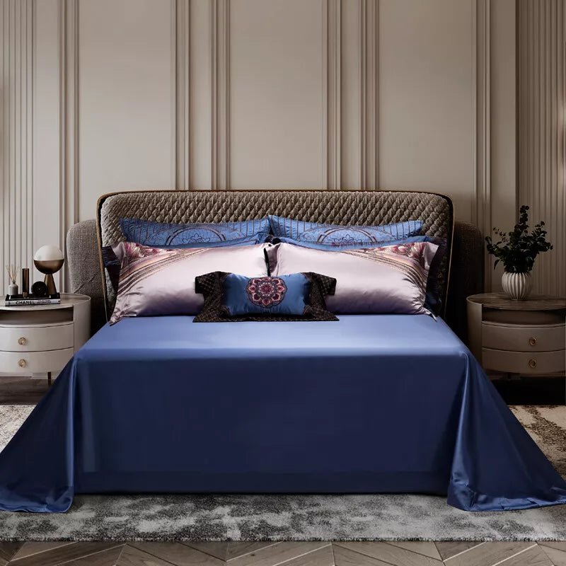 A neatly arranged bed featuring a blue satin bedspread with coordinating pillowcases in blue and lavender hues. The center pillow showcases a dark embroidered design with decorative ruffles. A quilted headboard and two side tables with lamps and a vase with greenery frame the setting, adding an elegant and modern ambiance.