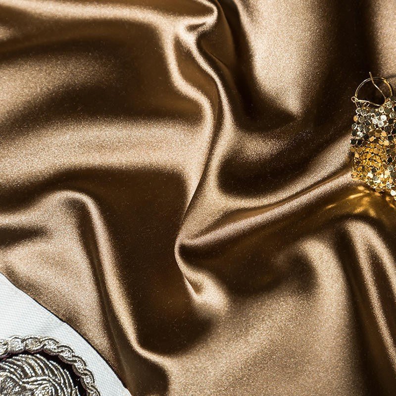 Smooth and lustrous gold fabric with a luxurious, silky texture that reflects light beautifully. The rich champagne hue adds a regal and elegant touch, accentuated by the subtle presence of decorative trim and golden embellishments. The material's sheen emphasizes its high-quality, premium finish.