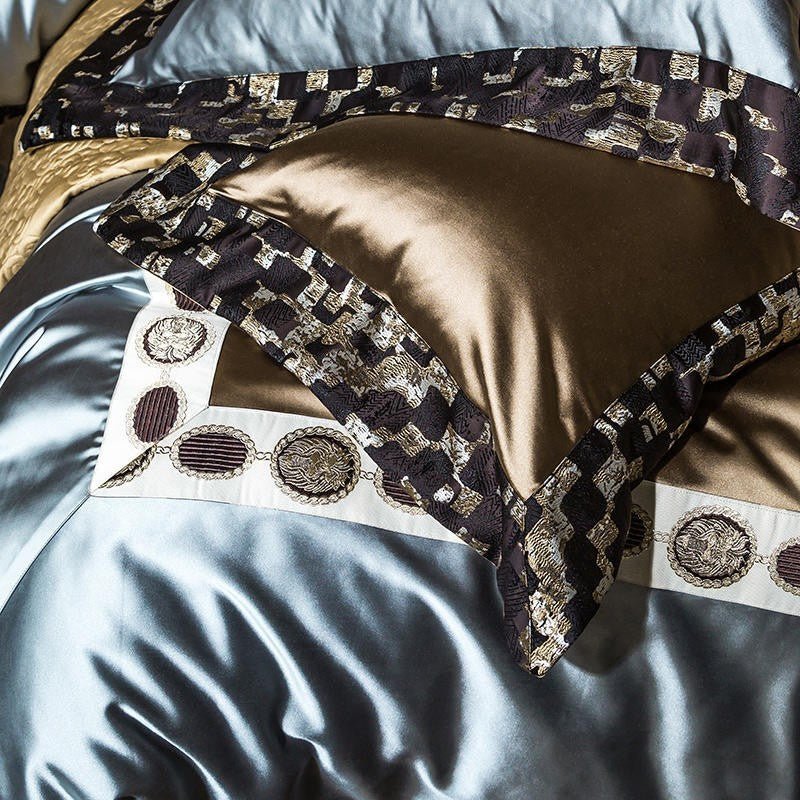 Close-up view of a bedding set showcasing intricate details and a combination of colors. The golden pillowcase features a smooth, silky texture with a subtle sheen, accented by a black and gold patterned border. The light blue satin fabric below reflects light with a soft luster, creating a luxurious contrast. Decorative trim with embroidered circular designs in varying shades of silver, black, and brown enhances the opulent appearance.